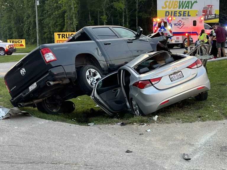 Black pickup truck on top of a silver car after a crash, with emergency responders on scene.